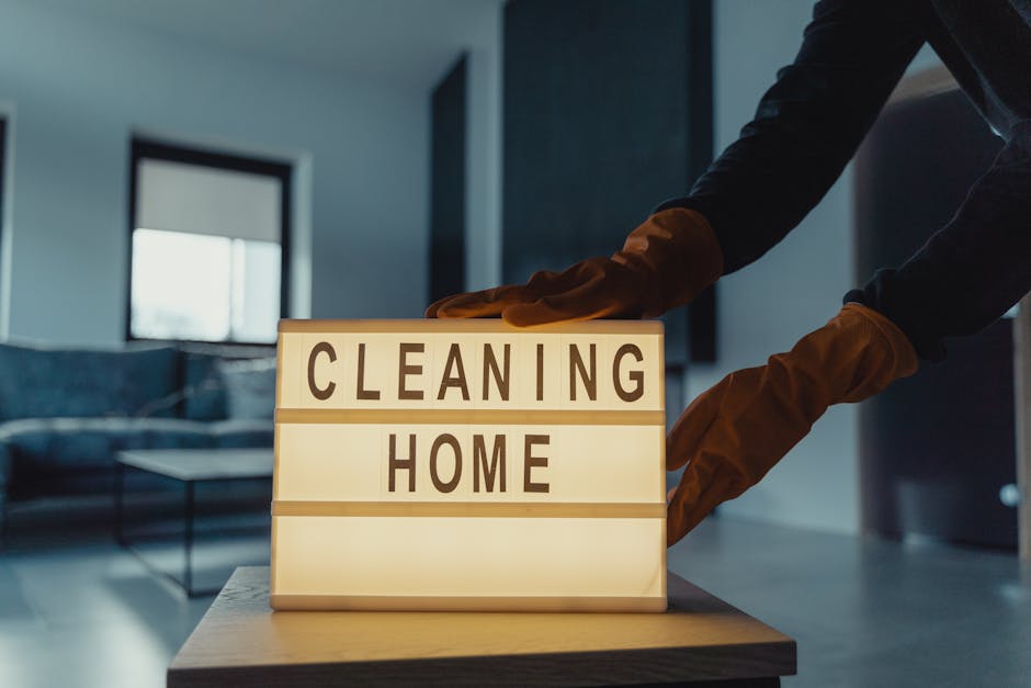 A commercial cleaning trolley positioned against a beige tiled wall in a maintenance or utility room, filled with various cleaning tools including mops, brooms, spray bottles, and cleaning cloths. To 