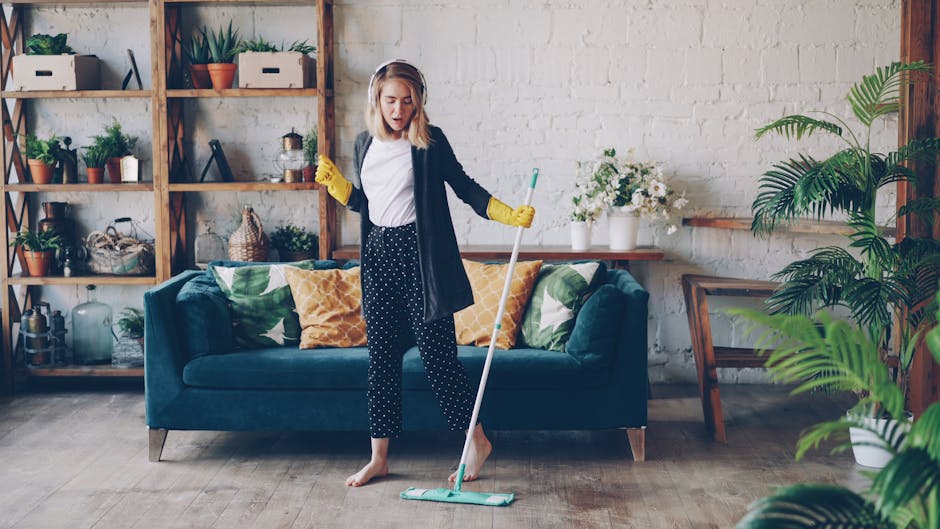 A woman engaged in domestic cleaning stands barefoot on a wooden floor in a modern living room, holding a mop with a light blue cleaning head. She is wearing yellow rubber gloves, a navy blazer, white shirt, and navy trousers with white polka dots. Behind her is a dark teal sofa adorned with patterned throw pillows in yellow, green, and white. To the left, there is a wooden shelving unit filled with potted plants, decorative jars, and books. The room features a white brick wall with a wooden picture frame leaning against it, and several potted green plants adding to the fresh, clean atmosphere. Bright, natural light illuminates the space, emphasizing the cleanliness of the surface areas as part of residential deep cleaning by Cleaners Ilford, focusing on surface care, hygiene, and overall maintenance.