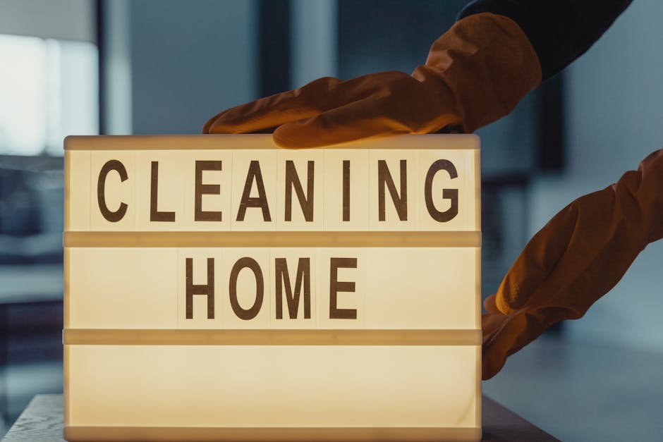 A person wearing orange cleaning gloves holding a lit-up sign with black letters spelling 'CLEANING HOME' on a lightbox, situated on a table in what appears to be a modern, well-lit interior space. The sign is illuminated, creating a warm glow around the text, symbolizing domestic cleaning and sanitisation processes. The background features blurred elements of a room, emphasizing the focus on the lightbox sign, which is associated with professional cleaning services by Cleaners Ilford. This setup suggests an emphasis on surface cleaning and maintaining hygiene in residential environments, aligned with the Ilford Broadway home cleaning guide for residents.