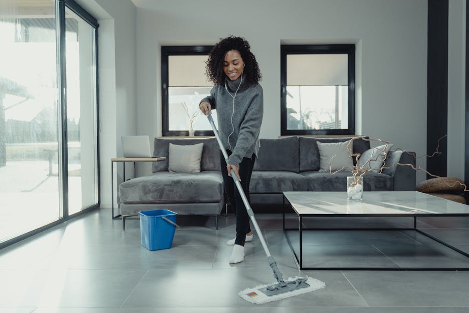 A woman engaged in domestic cleaning stands barefoot on a wooden floor in a modern living room, holding a mop with a light blue cleaning head. She is wearing yellow rubber gloves, a navy blazer, white shirt, and navy trousers with white polka dots. Behind her is a dark teal sofa adorned with patterned throw pillows in yellow, green, and white. To the left, there is a wooden shelving unit filled with potted plants, decorative jars, and books. The room features a white brick wall with a wooden picture frame leaning against it, and several potted green plants adding to the fresh, clean atmosphere. Bright, natural light illuminates the space, emphasizing the cleanliness of the surface areas as part of residential deep cleaning by Cleaners Ilford, focusing on surface care, hygiene, and overall maintenance.