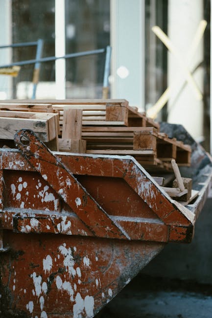 A man with curly hair, wearing a dark T-shirt and plaid shorts, stands on a sidewalk next to two large white construction waste bins filled with wooden planks, tiles, and debris. The scene is set outd